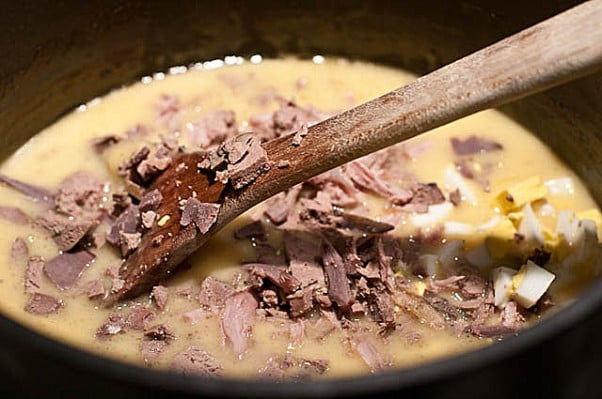 Image of cooked, chopped giblets and chopped eggs being added to gravy in a medium saucepan with a wooden spoon. // addapinch.com