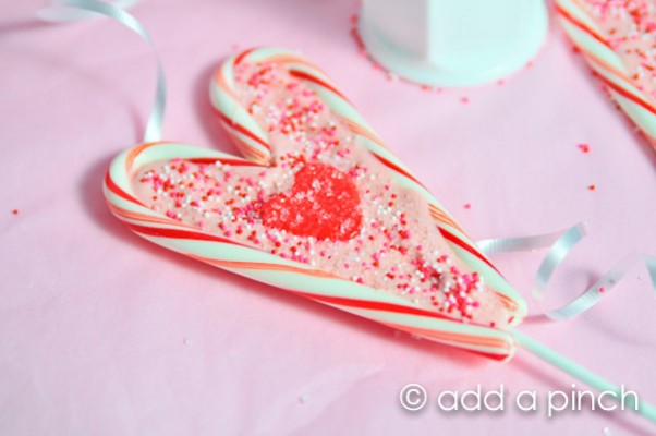 Heart shaped lollipop made of candy canes and melted candy melts sprinkled with nonpareils with a red heart frosting decoration in the center.