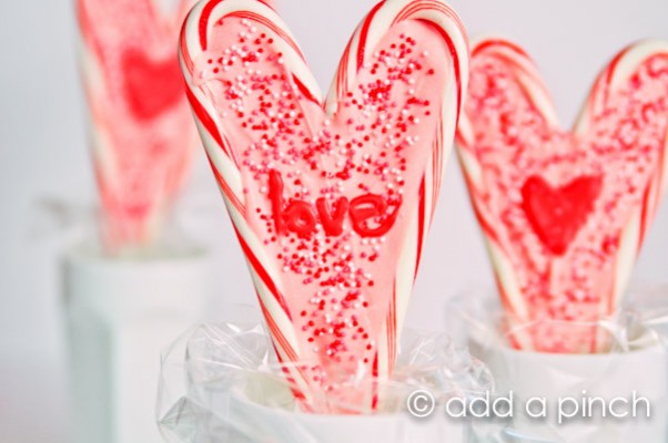 Three heart shaped lollipops made of candy canes and melted candy melts decorated with sprinkles and frosting.