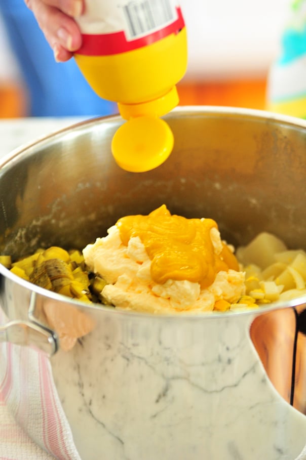 Adding yellow mustard to Potato salad ingredients of potatoes, diced eggs, chopped pickles, and mayonnaise in a large stainless pot. // addapinch.com