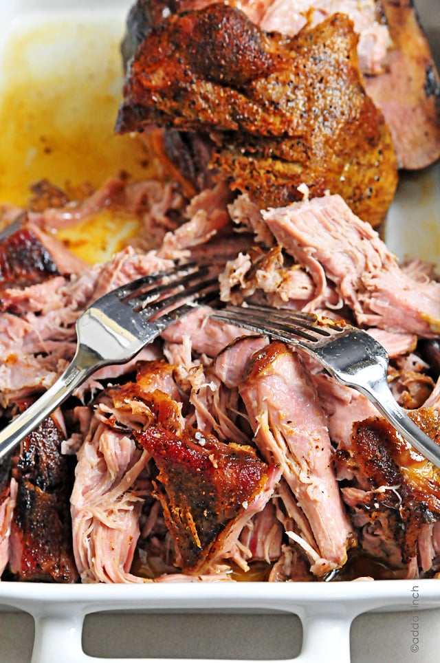 Two forks shred a tender, juicy pork roast on a white platter.