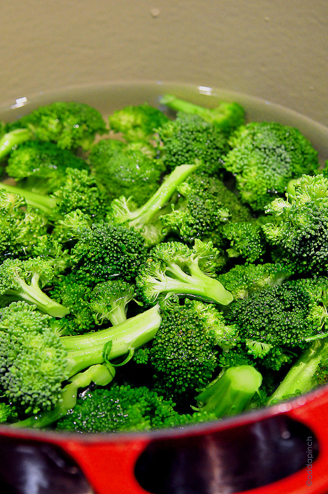 A bunch of fresh broccoli cut into florets is covered in water and ready to cook in a large red stockpot. 