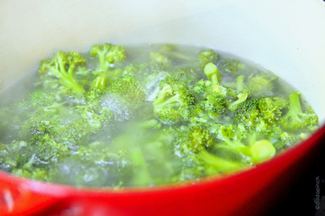 Red stockpot holds broccoli cooking in water on the stovetop. 