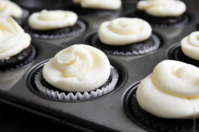 Chocolate cupcakes in a muffin pan topped with vanilla buttercream frosting.