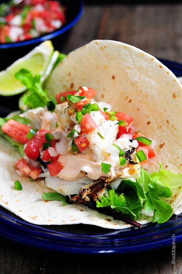 Fish taco topped with pico de gallo, salad greens and sriracha cream sauce, served on blue plate - addapinch.com