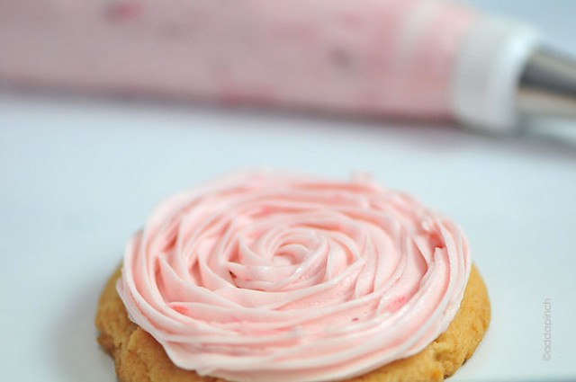 Sugar cookie covered in swirled topping of Strawberry Buttercream Frosting. Frosting bag with tip in background. 