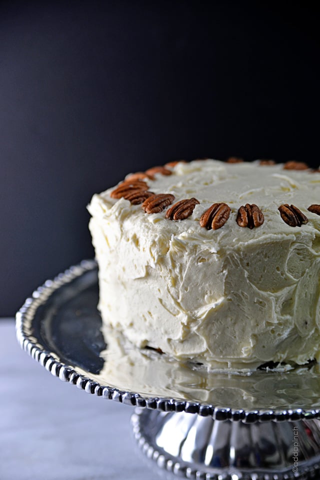 Cream Cheese frosted cake sits atop a silver toned cake stand. It is topped with pecans. 