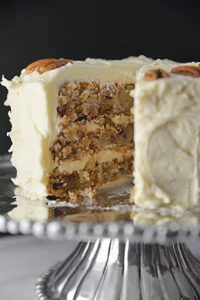 A slice is removed from a frosted Hummingbird Cake as it sits on a silver cake stand. 