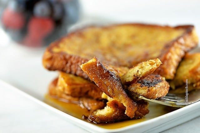 Horizontal photo of French Toast on a white plate with syrup and a bite on a silver fork. Mixed berries in the background.