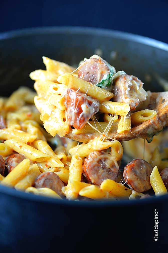Large pot of penne style pasta with red peppers flakes, slices of sausage, stringy melted cheese, and spinach all dipped with wooden spoon. 