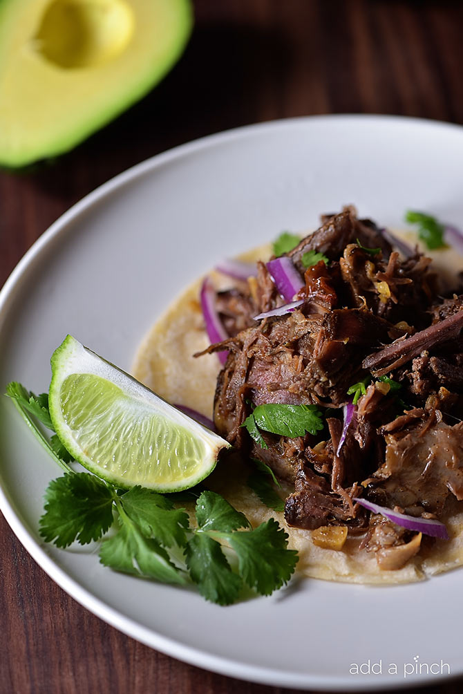 Photograph of tender barbacoa beef on a tortilla topped with red onions, cilantro, and served with lime. // addapinch.com