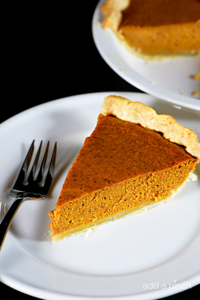 Slice of Pumpkin Pie on white plate with a fork and whole pie in the background 