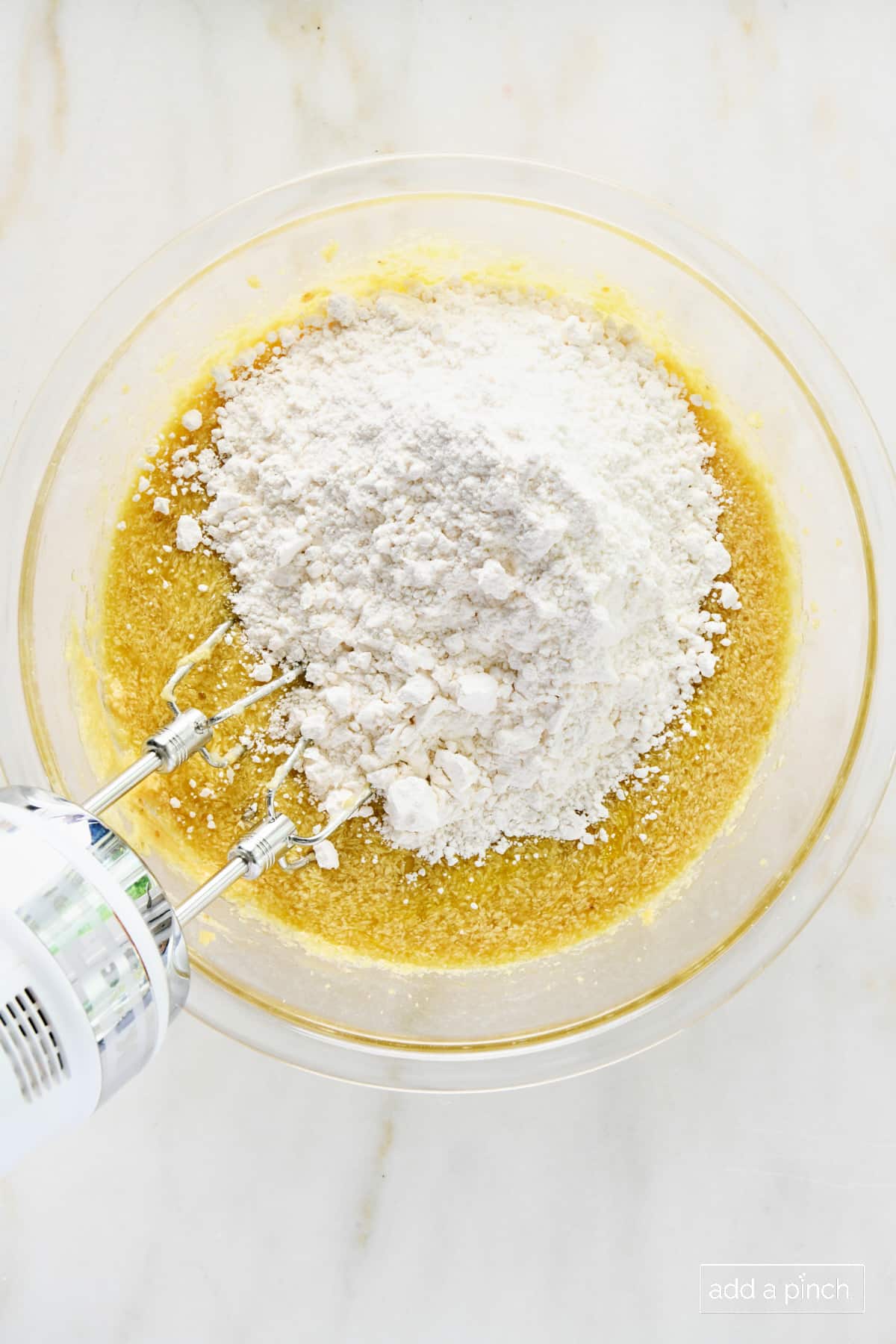 Flour added to batter in a glass bowl on a marble surface.