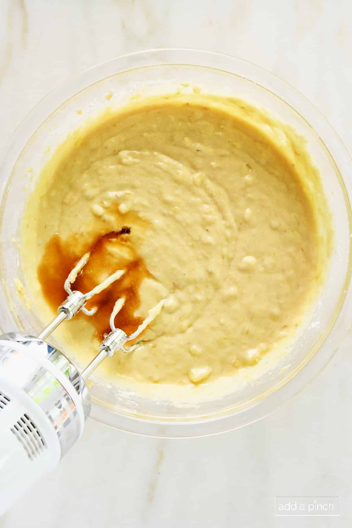 Vanilla extract being mixed into banana muffin batter in a glass bowl with a hand mixer.