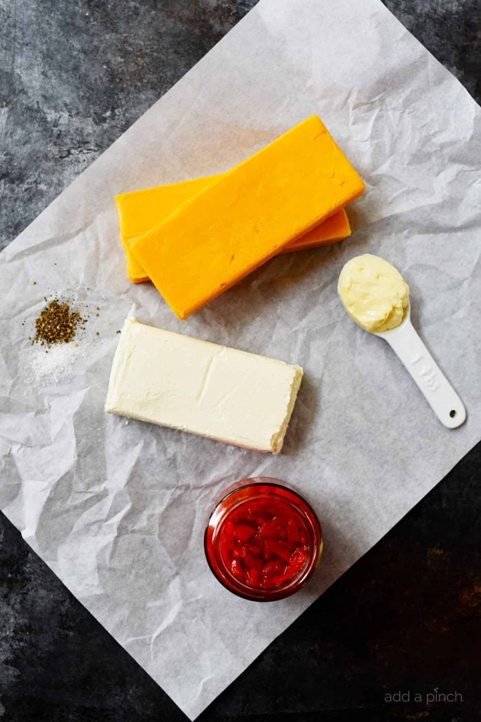 Ingredients for southern pimento cheese are placed on white baking paper - blocks of cheddar cheese and cream cheese, along with a spoon of mayonnaise and a glass container of pimentos and a scoop of seasoning blend. 