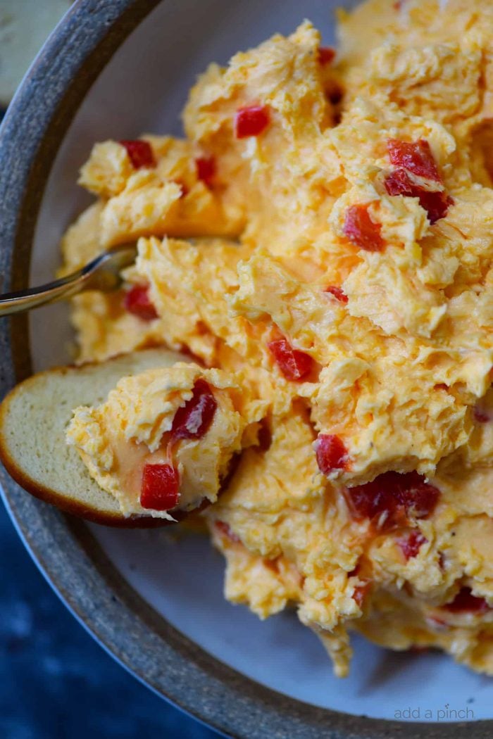 A bagel chip scoops up a bite of pimento cheese that's loaded with bits of pimento. It's served on a plate with a gray rim and a stainless fork. 