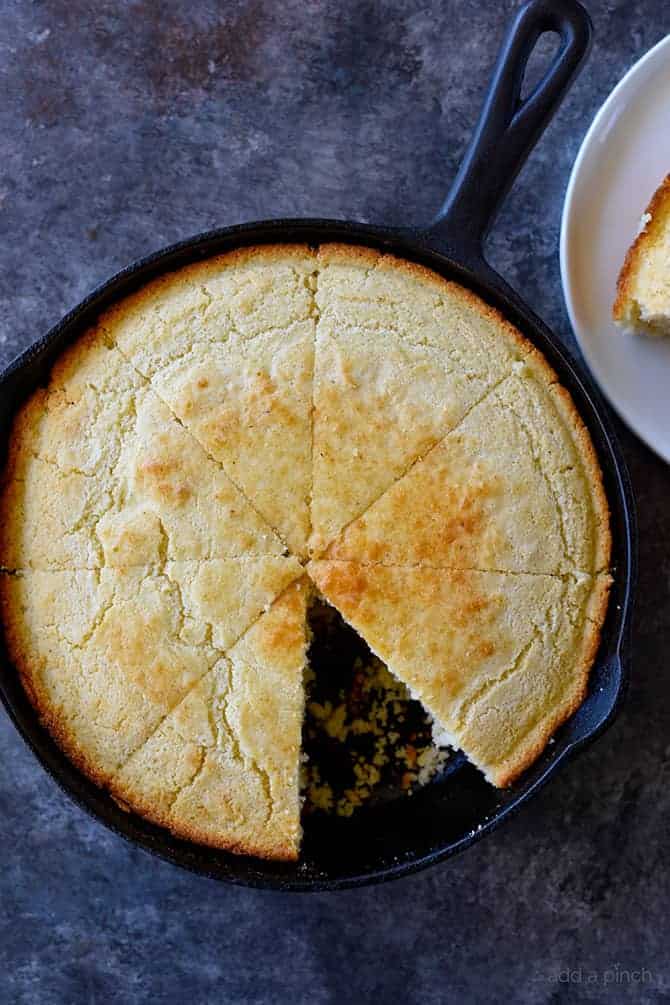 Cornbread cooked to a golden brown is sliced into wedges in a cast iron skillet with a slice missing. // addapinch.com