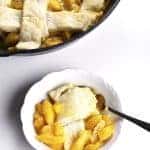 Skillet of lattice top peach cobbler with a serving in a white bowl nearby.