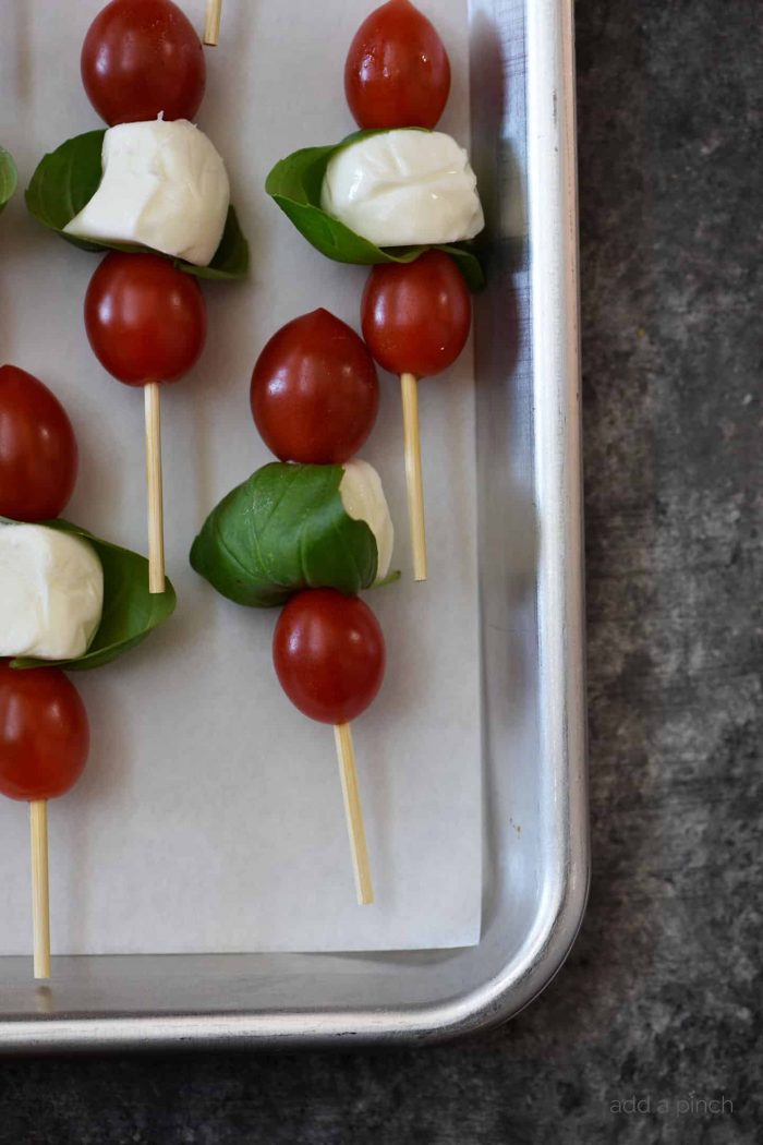 Baking sheet layerred with parchment holds bamboo skewered small tomatoes, mozzarella and basil // addapinch.com