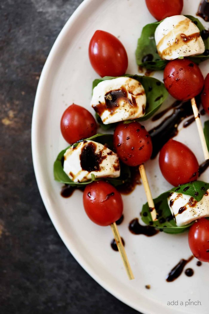 A white serving dish holds wooden party skewers layered with alternating small tomatoes, mozzarella balls and basil leaves. All are drizzled with balsamic glaze. // addapinch.com