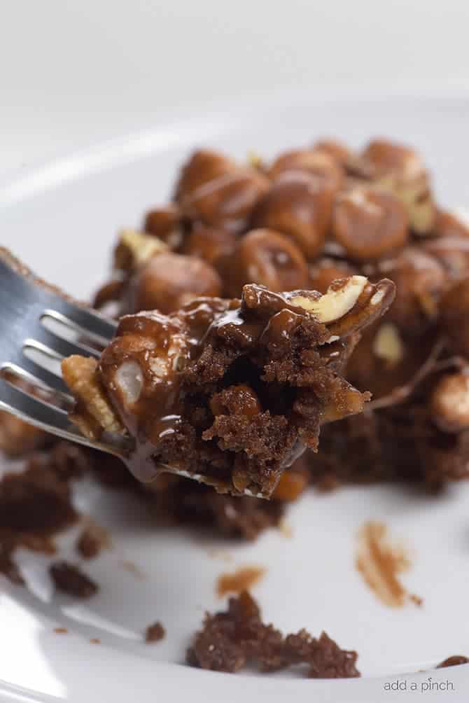 Bite of rich chocolate dessert topped with pecans, marshmallows and chocolate icing with remainder of serving on a white plate in the background. // addapinch.com