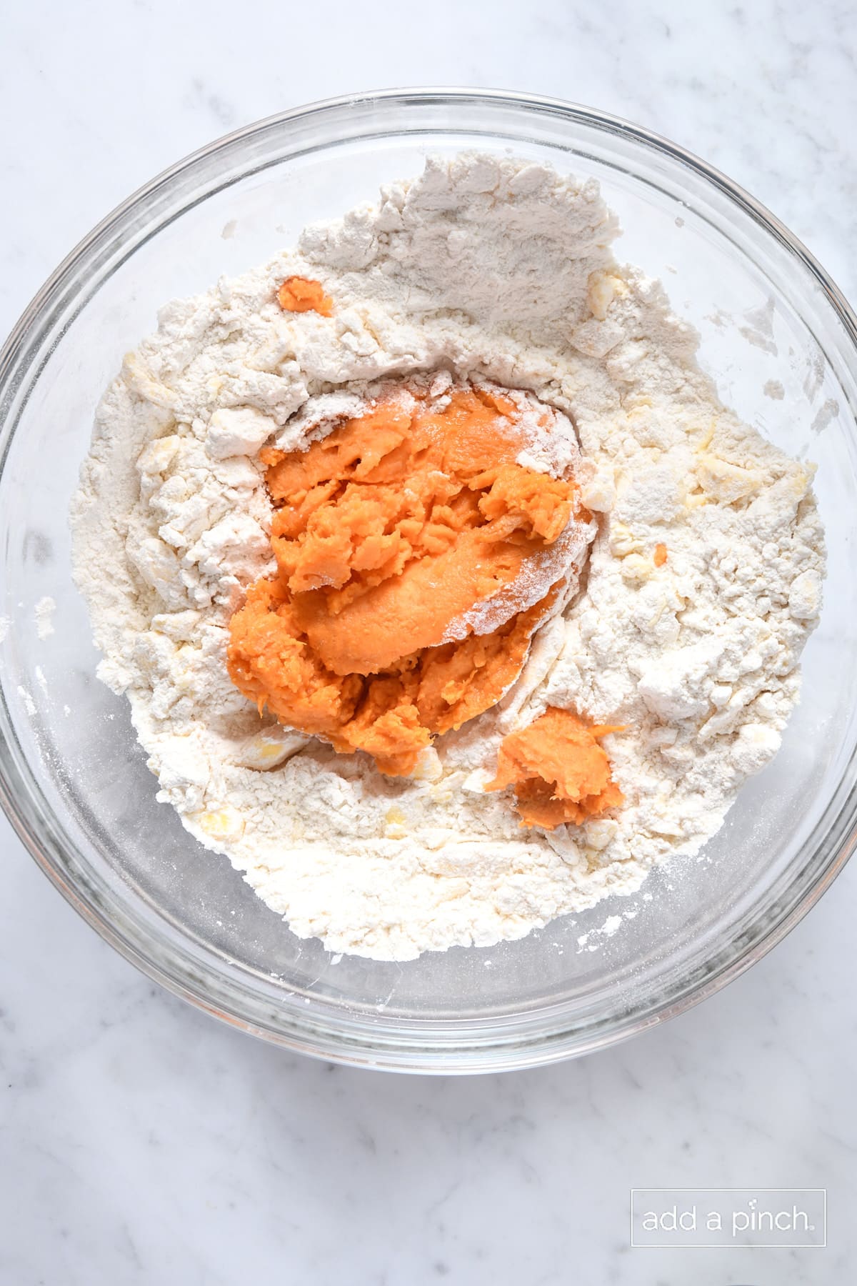 Mashed sweet potato added to flour mixture in a glass mixing bowl on a marble surface.
