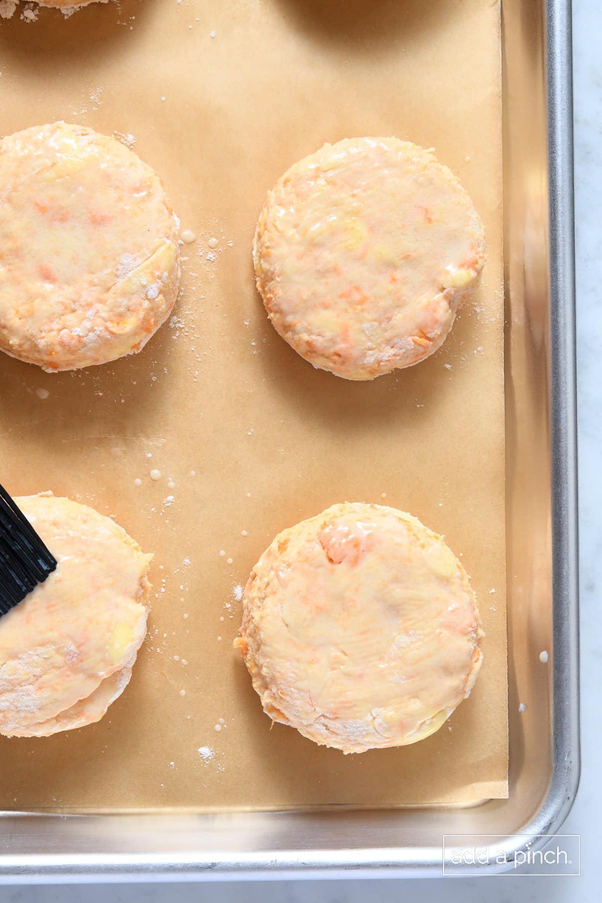 Brushing butter on sweet potato biscuits on a parchment lined baking sheet.