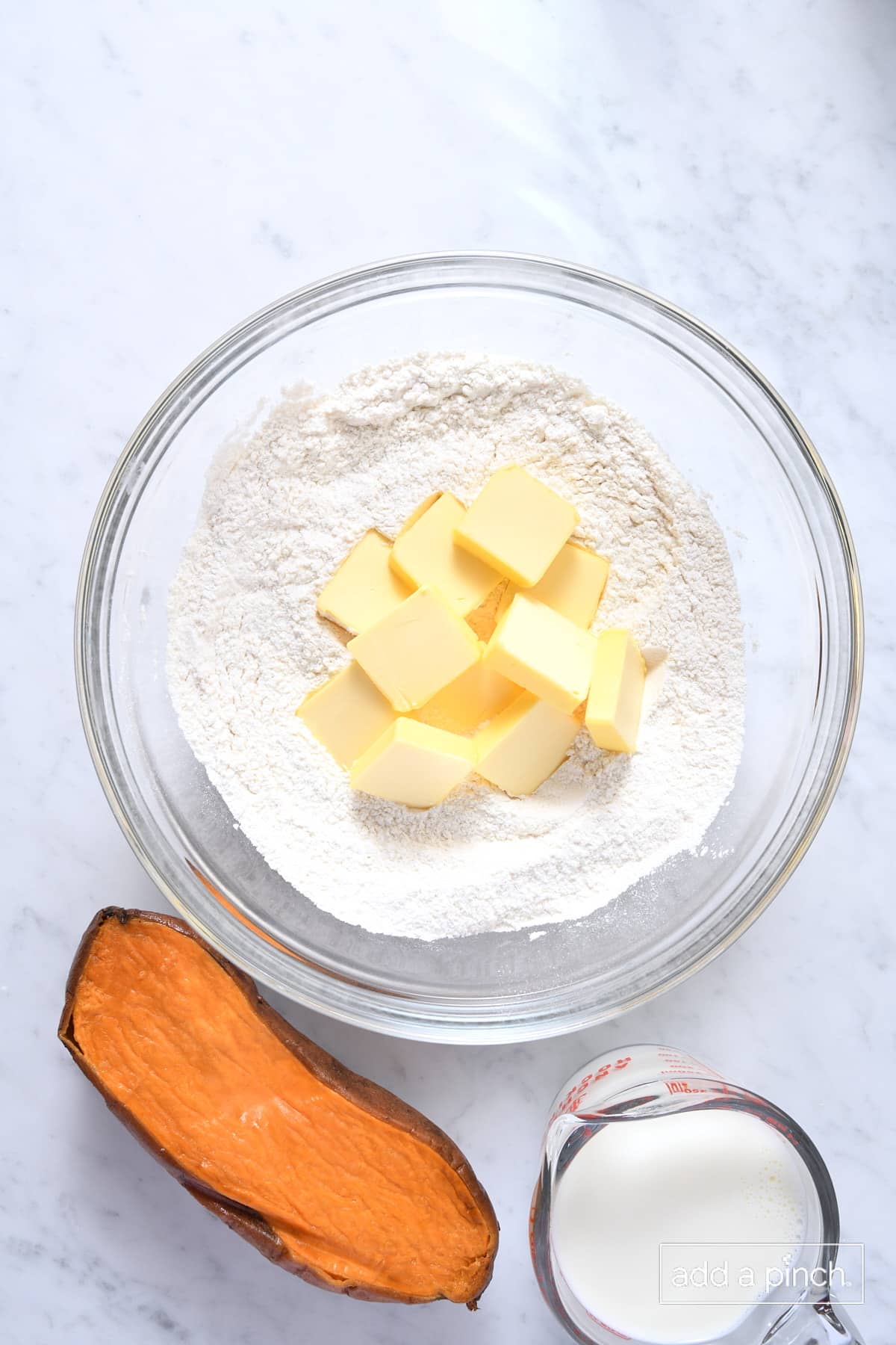 Butter added to glass bowl of flour with sweet potato and milk to the side on a marble counter.