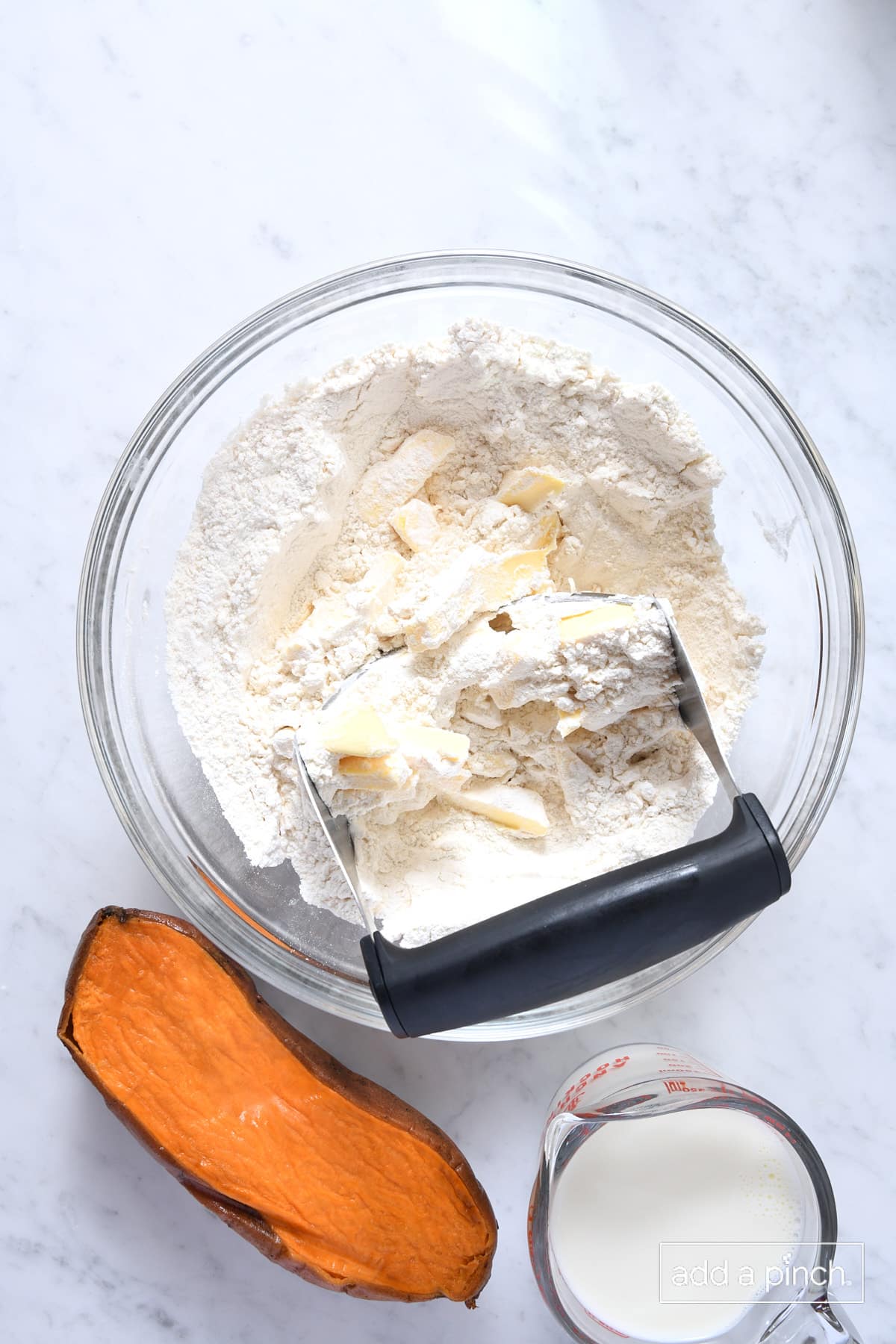 Pastry blender cutting butter into flour mixture in a glass bowl with sweet potato and milk to the side on a marble surface