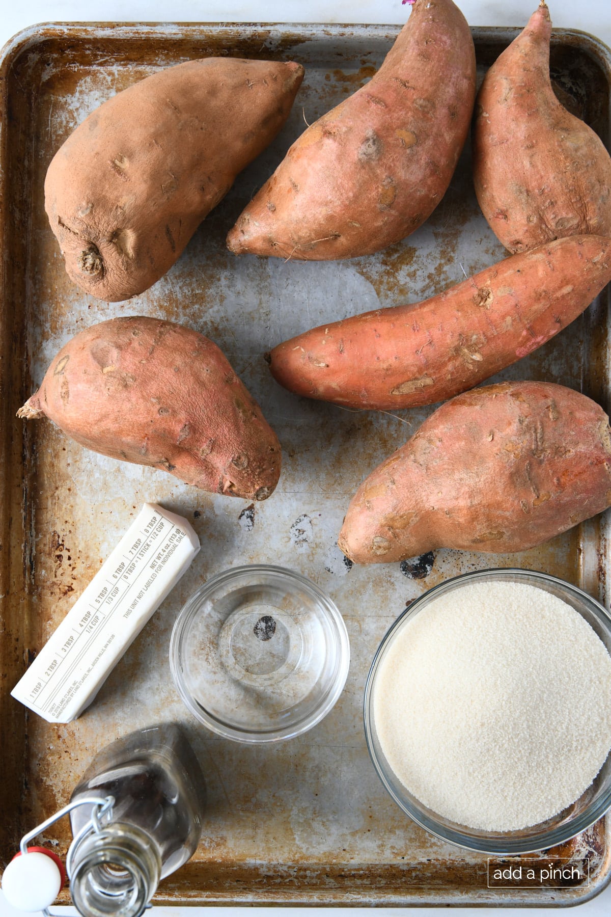 Ingredients used to make candied sweet potatoes on a baking sheet: sweet potatoes, butter, water, vanilla extract, and sugar.