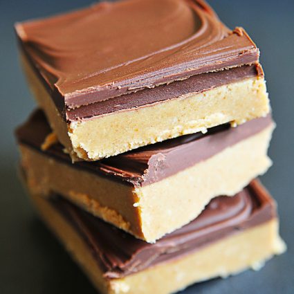 Photo of a stack of three peanut butter bars with a layer of peanut butter filling topped with a layer of chocolate on a dark background.