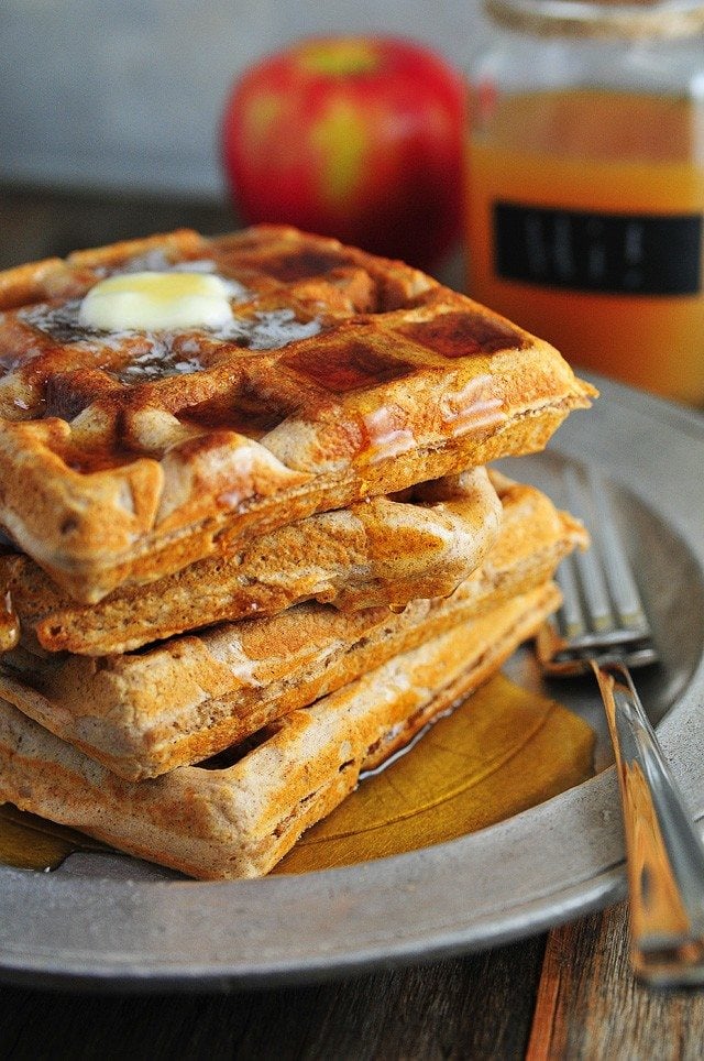 Stack of waffles on a pewter serving dish with fork. They are topped with melted butter and drizzled in maple syrup. Red apple and mug of apple cider in background // addapinch.com