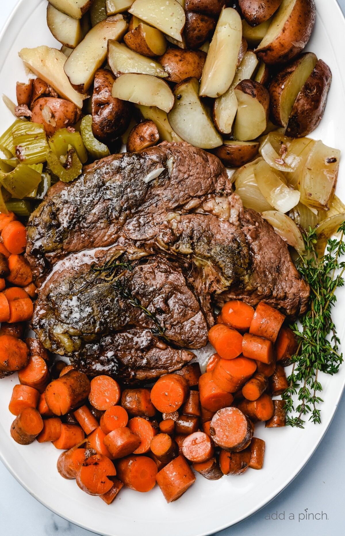 Photograph of cooked roast surrounded by potatoes, onions, fresh thyme, and carrots on a white platter. // addapinch.com