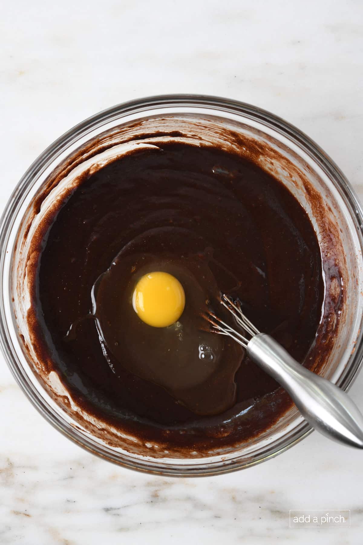 Egg added to chocolate mixture in a glass bowl with a metal whisk.