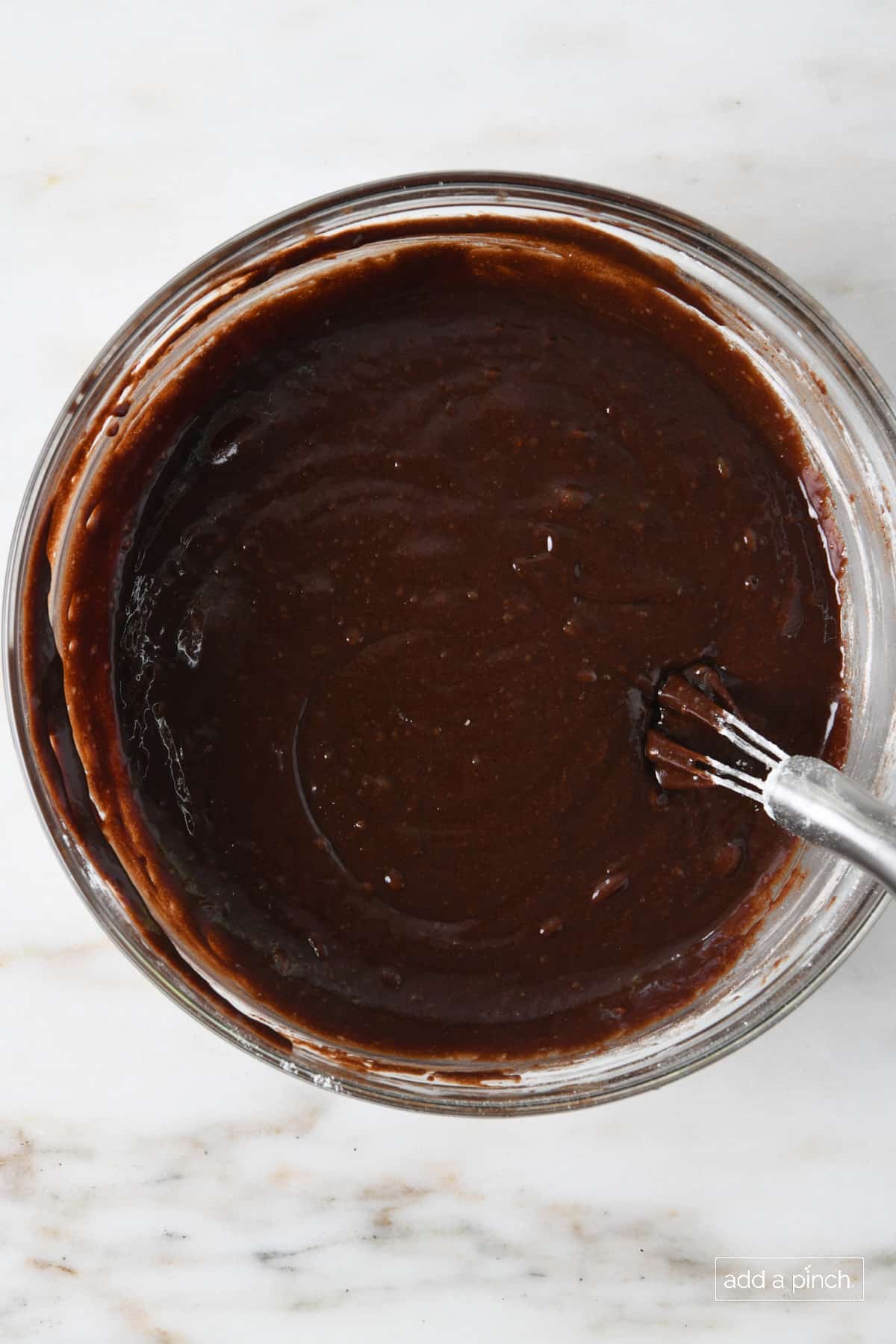 Thick, glossy chocolate brownie batter mixed in a glass bowl on marble counter, ready for add-ins.