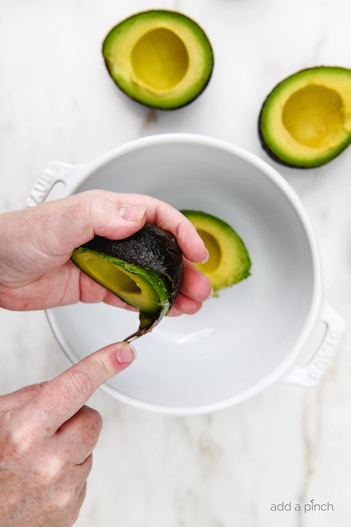 White bowl on marble countertop with  fresh avocado being scooped into it with a spoon. 