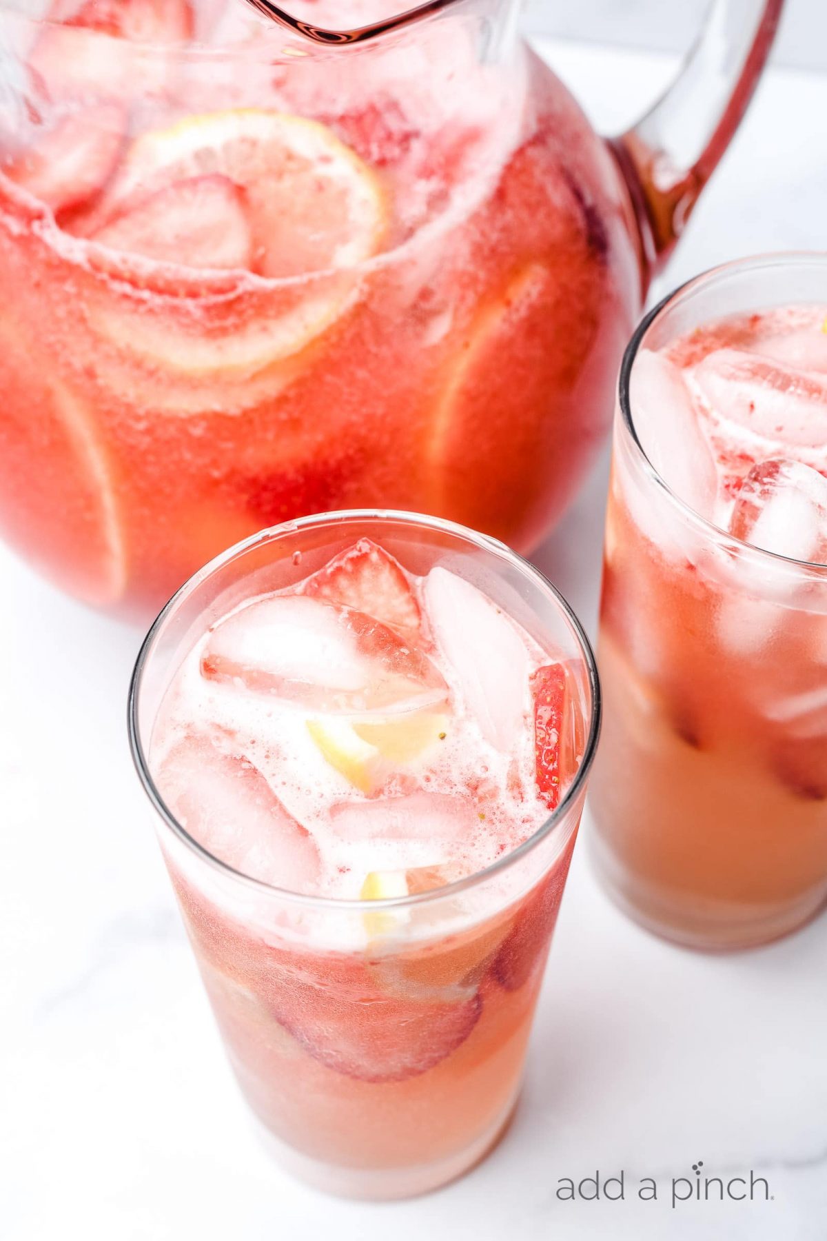 A pitcher of Strawberry Lemonade with strawberry and lemon slices sits beside two glasses with ice and full of the strawberry lemonade. 