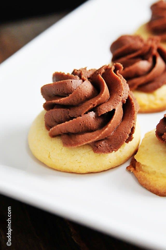 Golden butter cookies are decorated with swirls of chocolate buttercream frosting and served on a white tray.