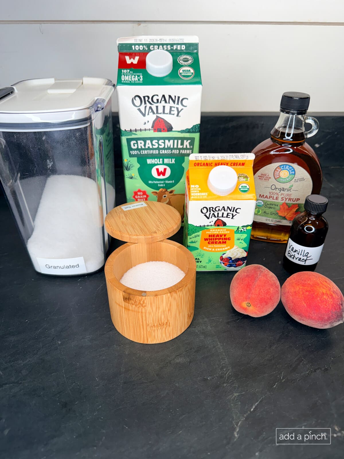 Ingredients for the best homemade peach ice cream recipe laid out on a marble counter: fresh ripe peaches, heavy whipping cream, whole milk, granulated sugar, pure maple syrup, and vanilla extract.