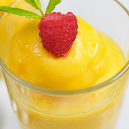 Frozen Peach Sorbet in a glass topped with fresh ripe raspberry and a sprig of mint on a white background.