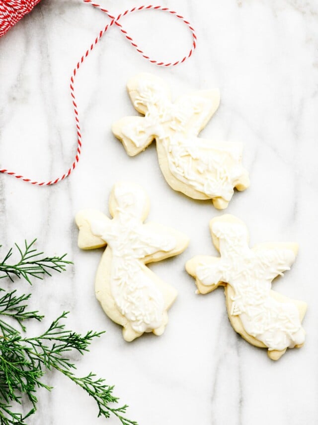 Marble countertop is decorated with iced angel shaped sugar cookies, surrounded by an evergreen twig and red and white twine.