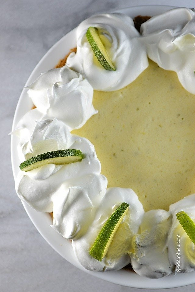 Photo of Key Lime Pie with whipped cream and lime wedges on a white pie plate.