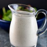 Image of homemade buttermilk ranch in a clear glass dressing jar. Salad in a blue bowl is in the background.