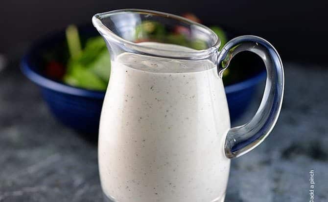 Image of homemade buttermilk ranch in a clear glass dressing jar. Salad in a blue bowl is in the background.