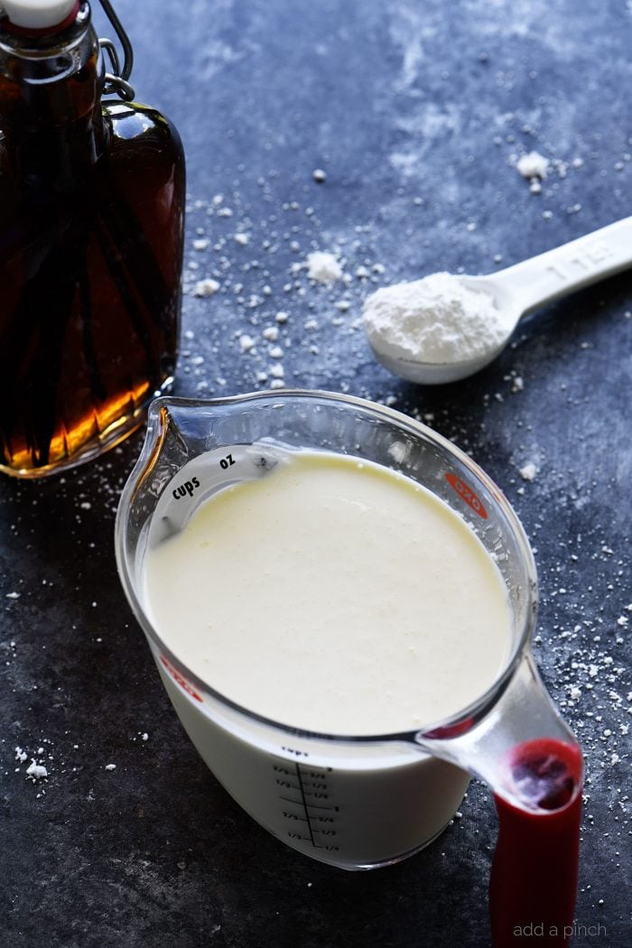 Ingredients for Whipped Cream - measuring cup of heavy whipping cream, glass bottle of Vanilla Extract and ceramic teaspoon of powdered sugar, all on stone counter sprinkled with some powdered sugar // addapinch.com