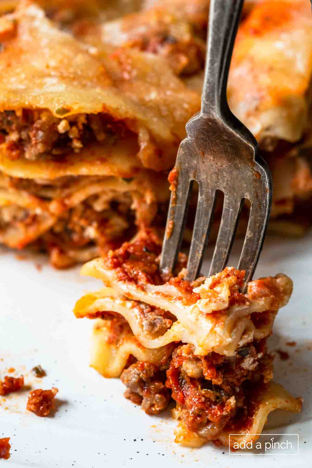 Photograph of a bite of cooked lasagna on a fork with piece of lasagna in the background.