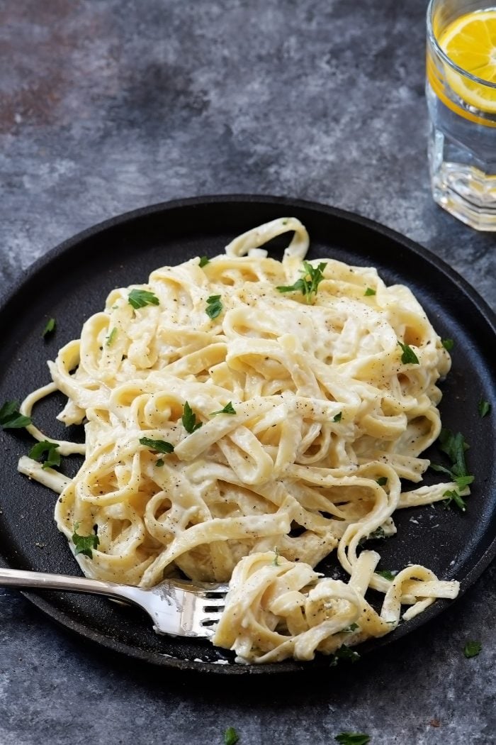 Fork with a twirl of Fettuccini Alfredo on a black plate, served with water with lemon wedge // addapinch.com