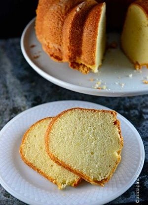 Image of two slices of pound cake on a white plate with the rest of the pound cake on a cake plate in the background.