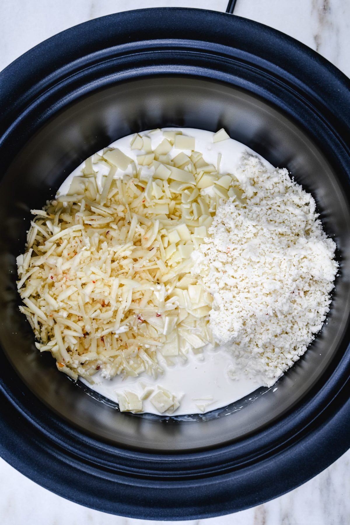 Grated cheeses and milk in a slow cooker insert to make queso.