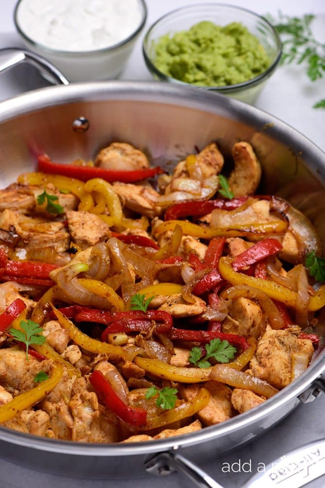 Skillet with chicken, red peppers, yellow peppers and onions, toped with cilantro sitting beside bowls of sour cream and guacamole // addapinch.com